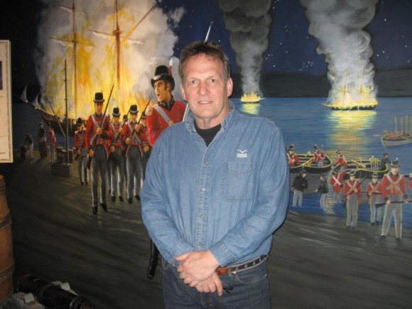 Executive Director Jerry Roberts in front of rendering of British burning American ships in Essex in 1814 